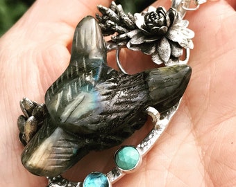 May include: A handcrafted silver pendant necklace featuring a carved labradorite bird. The pendant is adorned with silver floral accents and two small, round, blue and turquoise gemstones. The necklace is displayed against a blurred background.