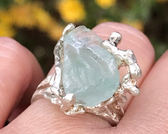 May include: A silver ring with a large, light blue aquamarine gemstone set in a textured, branch-like design.