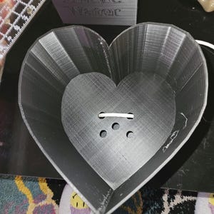 May include: A 3D-printed, heart-shaped planter in dark gray. The planter has a textured surface and a series of drainage holes at the bottom. A rectangular block with the words "Bitch Better Have Water" is visible in the background.