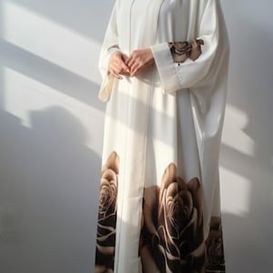 Peut inclure: Abaya blanc cassé avec une grande impression florale de roses marron. Le vêtement à manches longues présente un design fluide avec un motif floral assorti sur les manches et la partie inférieure. L'abaya est portée sur une robe assortie.