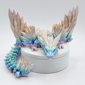May include: A fantasy dragon figurine with crystal-like wings and tail. The dragon is primarily white, with accents of blue, purple, and peach. It is posed on a white, round platform.