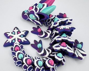 May include: A whimsical, articulated dragon figurine in shades of purple, white, pink, and teal. The dragon is made of multiple segments, with a snowflake-shaped ornament nearby. The dragon's head features pink and teal accents.