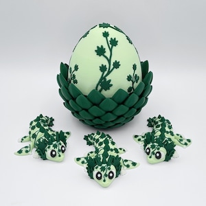 May include: A light green egg with dark green floral designs sits in a dark green nest. Three small, green and white dragon figurines are arranged in front of the egg, creating a fantasy theme.