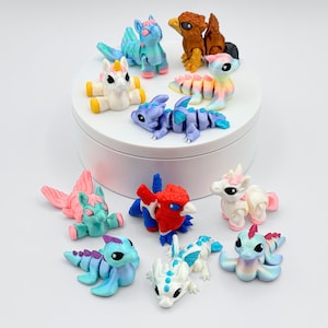 May include: A collection of colorful, miniature fantasy creatures, including dragons and unicorns, are arranged on a white surface. The figurines are made of clay and feature various colors like blue, pink, and white. Some are on a white platform.