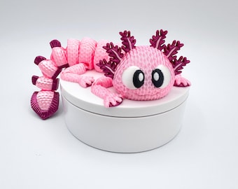 May include: A pink axolotl figurine with dark pink gills and accents, resting on a white circular base. The figurine has large, cartoonish eyes and a textured appearance, giving it a whimsical look. The axolotl's body is segmented, and its limbs are extended.