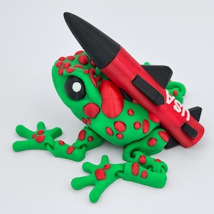 May include: A whimsical 3D-printed frog figurine in green with red spots, carrying a red and black rocket. The rocket has the word "USA" printed on it. The frog has large black eyes and outstretched limbs.