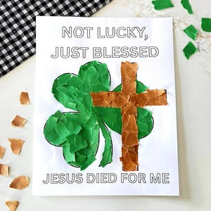 May include: A white paper with the text "NOT LUCKY, JUST BLESSED" and "JESUS DIED FOR ME". The paper features a green shamrock and a brown cross, both made of torn paper pieces. A black and white checkered cloth is in the background.
