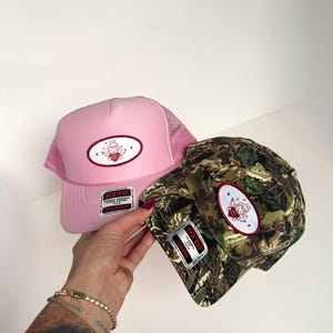 Puede incluir: Dos gorras de camionero. Una es rosa con una parte trasera de malla y un parche con un querubín con un corazón. La otra es de camuflaje con un parche de querubín similar. Ambas gorras tienen una pequeña etiqueta rectangular que dice "OTTO".