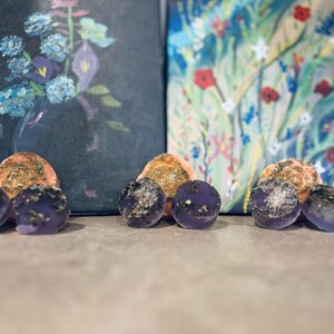 May include: Three purple and orange soap bars with a speckled texture. The soap bars are arranged in a row on a light brown surface.