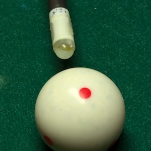 May include: A white billiard ball with a single red dot in the center. The ball is on a green felt surface.