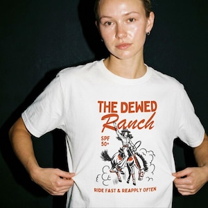 Pode incluir: Uma t-shirt branca de manga curta com uma impressão gráfica em vermelho e preto. O texto diz THE DEWED Ranch, SPF 50+ e RIDE FAST & REAPPLY OFTEN. A ilustração mostra uma pessoa a montar um cavalo com um chapéu de cowboy e botas.