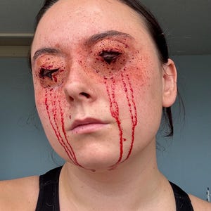 Puede incluir: Cara de una persona con maquillaje de efectos especiales que simula heridas abiertas alrededor de los ojos y sangre goteando por las mejillas. La piel es pálida con acentos rojos, creando un efecto dramático y sangriento. La persona lleva una camiseta sin mangas negra.