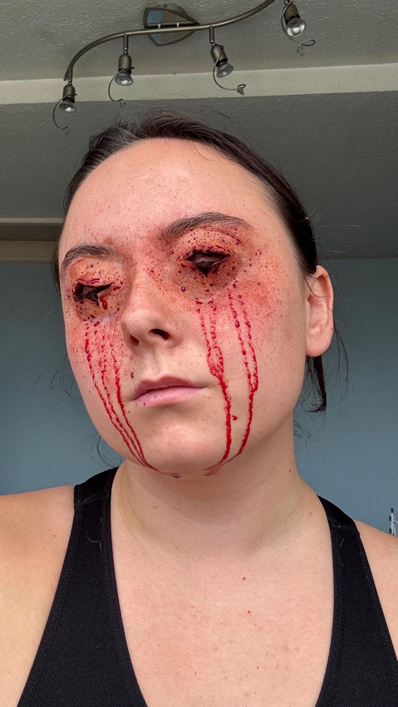 Puede incluir: Cara de una persona con maquillaje de efectos especiales que simula heridas abiertas alrededor de los ojos y sangre goteando por las mejillas. La piel es p&aacute;lida con acentos rojos, creando un efecto dram&aacute;tico y sangriento. La persona lleva una camiseta sin mangas negra.