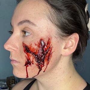 Puede incluir: Vista de perfil de un rostro con maquillaje de efectos especiales. El maquillaje representa una herida profunda y abierta con tejido expuesto y sangre goteando. El efecto es detallado y dramático, con tonos rojos oscuros y marrones.