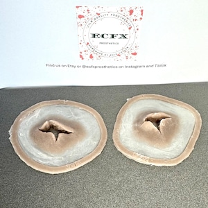 Puede incluir: Dos pr&oacute;tesis de heridas. Cada una tiene un centro marr&oacute;n claro con una hendidura, rodeado por un &oacute;valo blanco y marr&oacute;n claro. El texto "ECFX PROSTHETICS" es visible en el fondo.