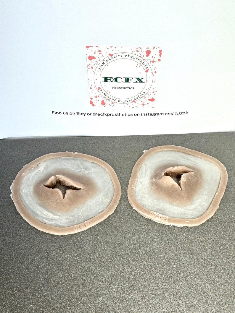 Puede incluir: Dos pr&oacute;tesis de heridas. Cada una tiene un centro marr&oacute;n claro con una hendidura, rodeado por un &oacute;valo blanco y marr&oacute;n claro. El texto "ECFX PROSTHETICS" es visible en el fondo.