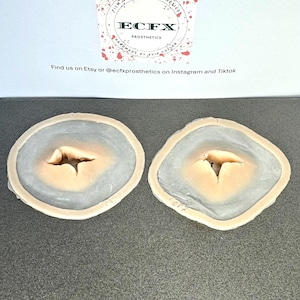 Puede incluir: Dos pr&oacute;tesis de heridas. Cada una tiene un centro de color melocot&oacute;n claro con un anillo exterior gris-azulado. La parte superior de la imagen incluye un logotipo que dice "ECFX PROSTHETICS" y "FILM QUALITY PROSTHETICS".