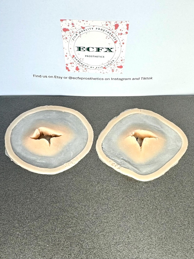 Puede incluir: Dos pr&oacute;tesis de heridas. Cada una tiene un centro de color melocot&oacute;n claro con un anillo exterior gris-azulado. La parte superior de la imagen incluye un logotipo que dice "ECFX PROSTHETICS" y "FILM QUALITY PROSTHETICS".