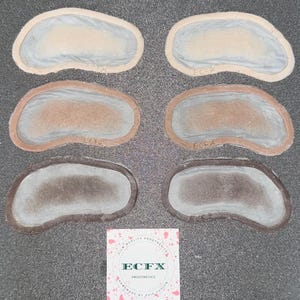 Pode incluir: Seis peças protéticas em tons de pele variados, dispostas em duas filas de três. Cada peça tem uma forma arredondada com uma superfície texturizada. Um pequeno cartão quadrado com o texto "ECFX PROSTHETICS" na parte inferior.