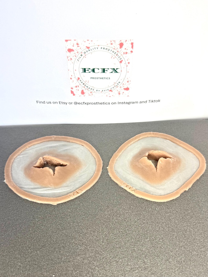 Puede incluir: Dos pr&oacute;tesis de heridas. Cada una tiene un centro color carne con una abertura profunda e irregular, rodeada de un material transl&uacute;cido similar a la piel y un borde fino color carne. El logotipo de ECFX es visible.