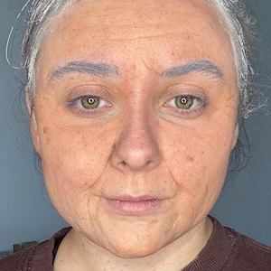 Może przedstawiać: Zbliżenie portretu osoby z makijażem nałożonym tak, aby przypominał osobę starszą. Odcień skóry to mieszanka pomarańczowego i brązowego, ze zmarszczkami i plamami starczymi. Osoba ma szare brwi i włosy i ma na sobie brązowy sweter.