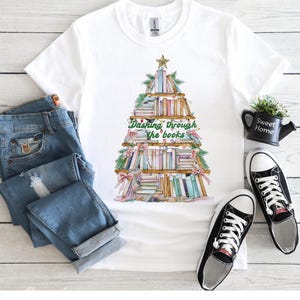 May include: White t-shirt with a Christmas tree design made of books. The tree is decorated with green garland, red bows, and a gold star. The text "Dashing through the books" is printed below the tree.