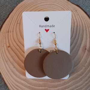 May include: A pair of brown, round, handmade earrings with gold hooks. The earrings are displayed on a white card with the word "Handmade" and a red heart.
