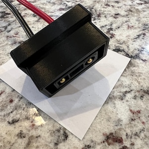 Pode incluir: Conector de plástico preto com dois pinos dourados. O conector está ligado a dois fios, um preto e um vermelho.