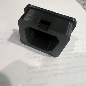 Peut inclure: Un objet en plastique noir imprimé en 3D avec une base carrée et une ouverture hexagonale. L'objet est conçu pour s'adapter à un article spécifique, tel qu'un téléphone ou un petit appareil.