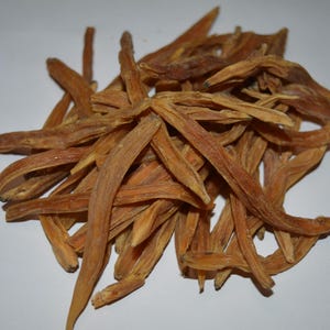 May include: A pile of dried, light brown, elongated roots. The roots have a slightly curved shape and a rough texture. They are arranged in a loose heap against a white background. The color is consistent throughout the pile.