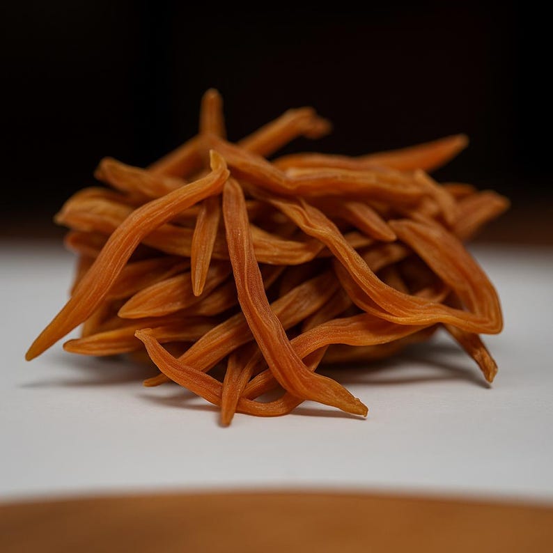 May include: A pile of dried, orange-brown, slender, curved objects, possibly a spice or herb, against a white surface. The objects have a textured appearance and are clustered together, with some extending outwards. The background is dark.