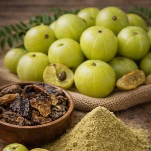 Könnte beinhalten: Ein Stillleben mit frischen grünen Amla-Früchten, getrockneten Amla-Stücken in einer Holzschale und einem Haufen Amla-Pulver. Die Früchte sind ganz und halbiert und zeigen ihr Inneres. Auf einer rustikalen Holzoberfläche.
