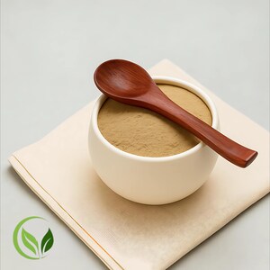 May include: A white ceramic bowl filled with a light brown powder, topped with a dark wooden spoon. The bowl rests on a beige napkin, with a green leaf logo in the corner. The spoon is approximately 15 cm long.