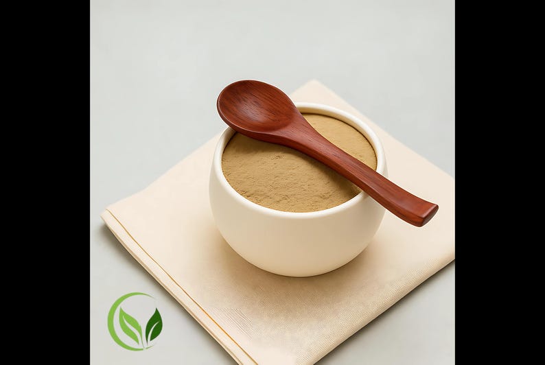 May include: A white ceramic bowl filled with a light brown powder, topped with a dark wooden spoon. The bowl rests on a beige napkin, with a green leaf logo in the corner. The spoon is approximately 15 cm long.