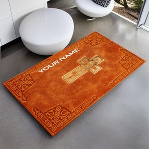 Puede incluir: Una alfombra de entrada de cuero marrón con un borde de nudo celta y una cruz dorada con una figura de Jesucristo. El texto "YOUR NAME" está impreso en blanco en la alfombra.