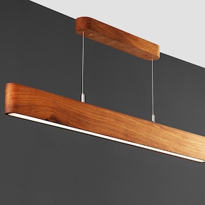 May include: A modern wooden pendant light fixture with a long, rectangular shape. The light is suspended from the ceiling by two thin wires.
