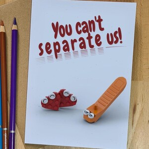 May include: A white greeting card with the red text "You can't separate us!" featuring a graphic of red and orange plastic building blocks with cartoon eyes. Beside the card are five coloured pencils in yellow, black, blue, brown, and purple.