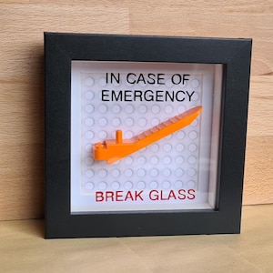 May include: A square black shadow box frame containing a white building block baseplate. Mounted on the centre is an orange plastic brick separator tool. The text IN CASE OF EMERGENCY is printed in black above, and BREAK GLASS in red below.