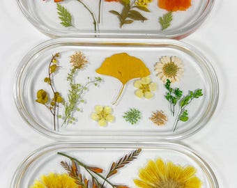 May include: Three oval-shaped clear resin trays with pressed flowers and leaves. The top tray features orange and yellow flowers, the middle tray features yellow and green leaves and flowers, and the bottom tray features yellow flowers.