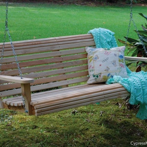 May include: A wooden swing bench with a light brown finish hangs from chains. A light blue crocheted blanket and a white pillow with a floral pattern are on the bench. The swing is in a grassy area with green foliage in the background.