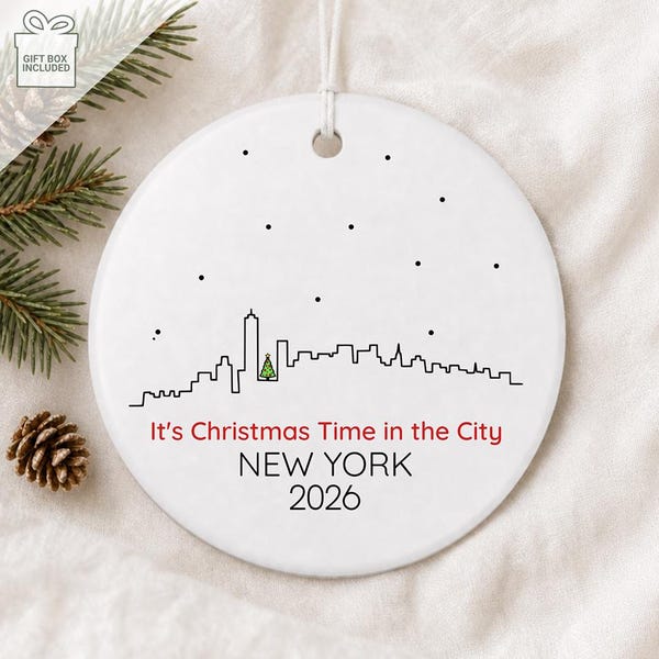May include: A round white ceramic ornament featuring a black line art skyline of New York City with a small green Christmas tree. The text reads It's Christmas Time in the City, NEW YORK, and 2026 in black and red lettering with black dots.