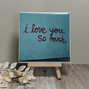 May include: A square ceramic tile with the handwritten phrase "i love you So much" in red script on a teal background. The tile is displayed on a small wooden easel, with a sprig of white flowers in the foreground.