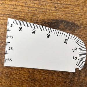 May include: White protractor with black markings, showing degree measurements from 5 to 60 degrees. The protractor is made of plastic and has a rounded edge.