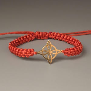 May include: A vibrant orange macrame bracelet featuring a gold-colored, four-pointed star charm. The adjustable bracelet is made of woven cord and is displayed against a neutral gray background. The bracelet is a piece of jewelry.