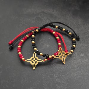 May include: Two beaded bracelets. One bracelet features red and gold beads with a gold-colored charm. The other bracelet has black and gold beads with a gold-colored charm. Both bracelets have adjustable red and black cords.