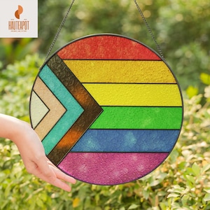 Op de afbeelding: Een rond gebrandschilderd glazen paneel met een regenboogkleurenschema en een geometrisch ontwerp. Het paneel is opgehangen aan een ketting en bevat de tekst "HAUTEAPOT". De kleuren zijn rood, geel, groen, blauw en roze.