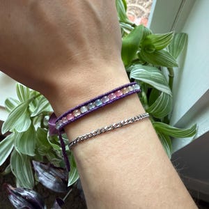 May include: A close-up of a wrist adorned with two bracelets. The top bracelet is purple with a row of colorful beads. Below it is a silver chain bracelet. The bracelets are displayed against a background of green plants and a window.