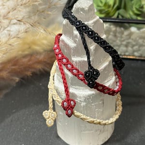 Op de afbeelding: Drie macramé armbanden in zwart, rood en crème worden getoond. De armbanden zijn versierd met kleine kralen en ingewikkelde knopen. De armbanden zijn gestapeld op een wit, getextureerd cilindrisch object, dat het vakmanschap laat zien.