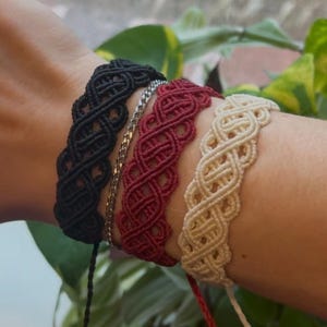 May include: Close-up of three macrame bracelets in black, burgundy, and cream colors. The bracelets have a woven design and are displayed on a wrist. A silver chain bracelet is also visible. The background includes green foliage.