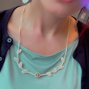 May include: A cream-colored, crocheted necklace with a leaf design and a central, round, amber-colored bead. The necklace is worn over a light blue top. Silver earrings with white accents are also visible.